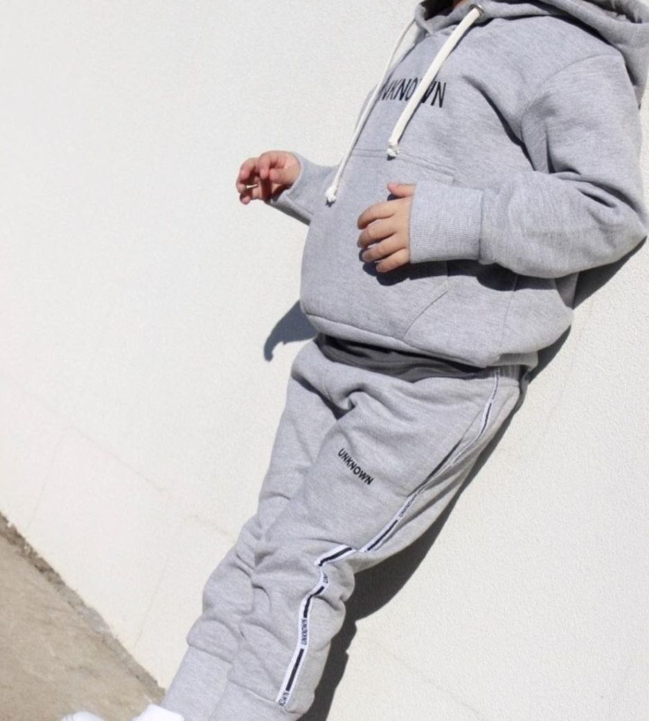 Young boy wearing THE UNKNOWN CHILDREN Originals Grey Tracksuit Casual Look, standing against a white wall