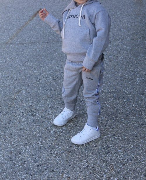 Child wearing a gray THE UNKNOWN CHILDREN tracksuit with white sneakers on a concrete surface. The pants feature a sleek design, perfect for active play and casual wear.