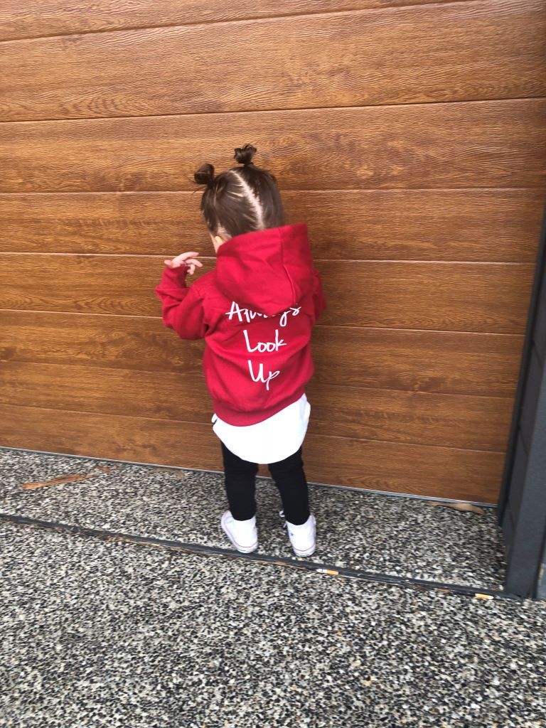 A young child wearing THE UNKNOWN CHILDREN 'Originals' "Always Look Up" red hoodie stands against a wooden wall. The unisex, limited-edition hoodie is made from 100% superior cotton, offering a trendy and comfortable style for boys and girls.