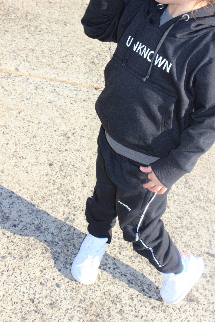 A child wearing a black "UNKNOWN" hoodie paired with matching tracksuit pants and white sneakers. The outfit offers a sleek, comfortable, and stylish look, ideal for both casual wear and active play.