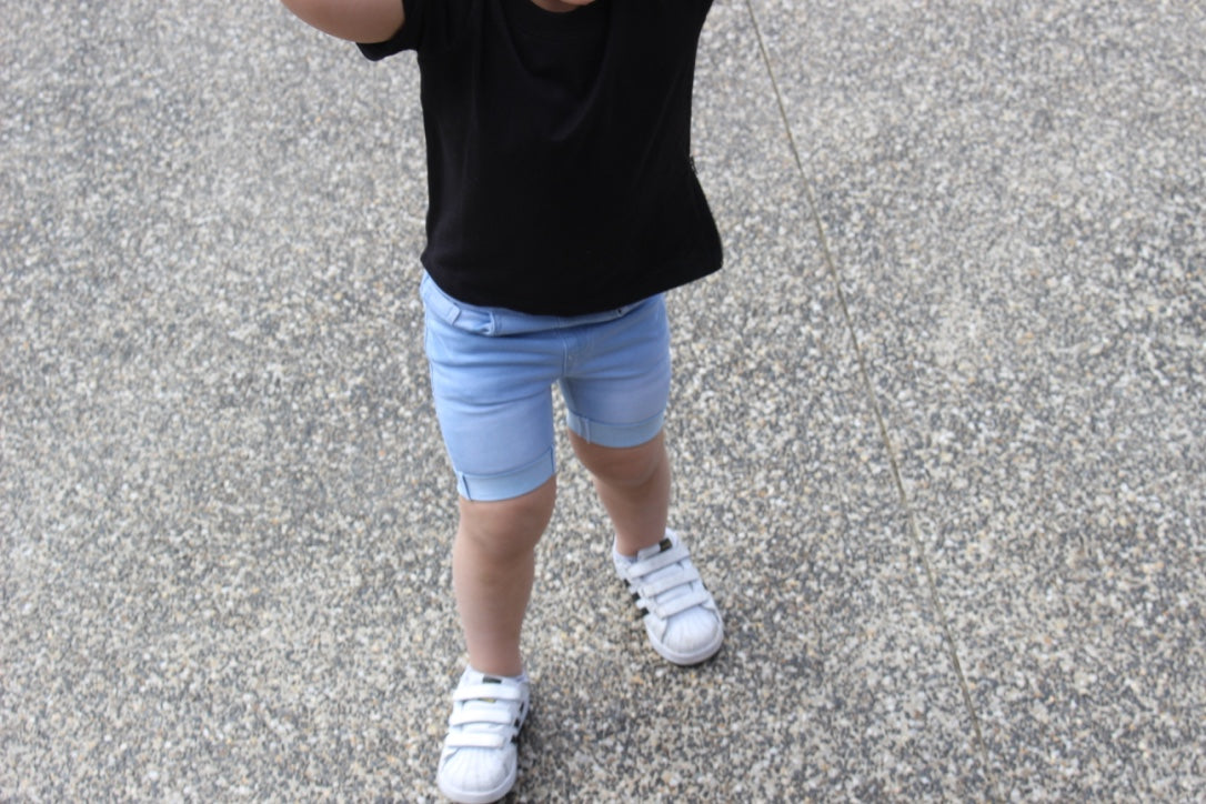Child wearing a black t-shirt paired with light blue rolled hem denim shorts and white sneakers. The outfit offers a casual and stylish look for active play or everyday wear.