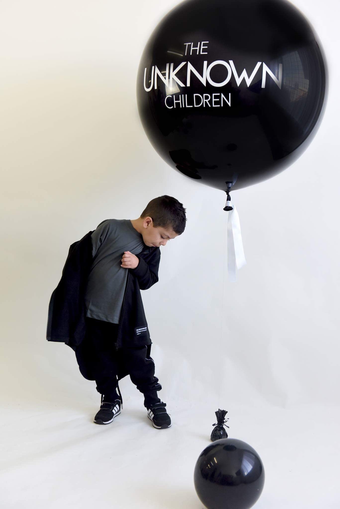 Child wearing a gray t-shirt and black tracksuit pants with "UNKNOWN" branding, adjusting their black zip-up trench. A large black balloon with "THE UNKNOWN CHILDREN" logo is floating beside them, with smaller black balloons on the floor.