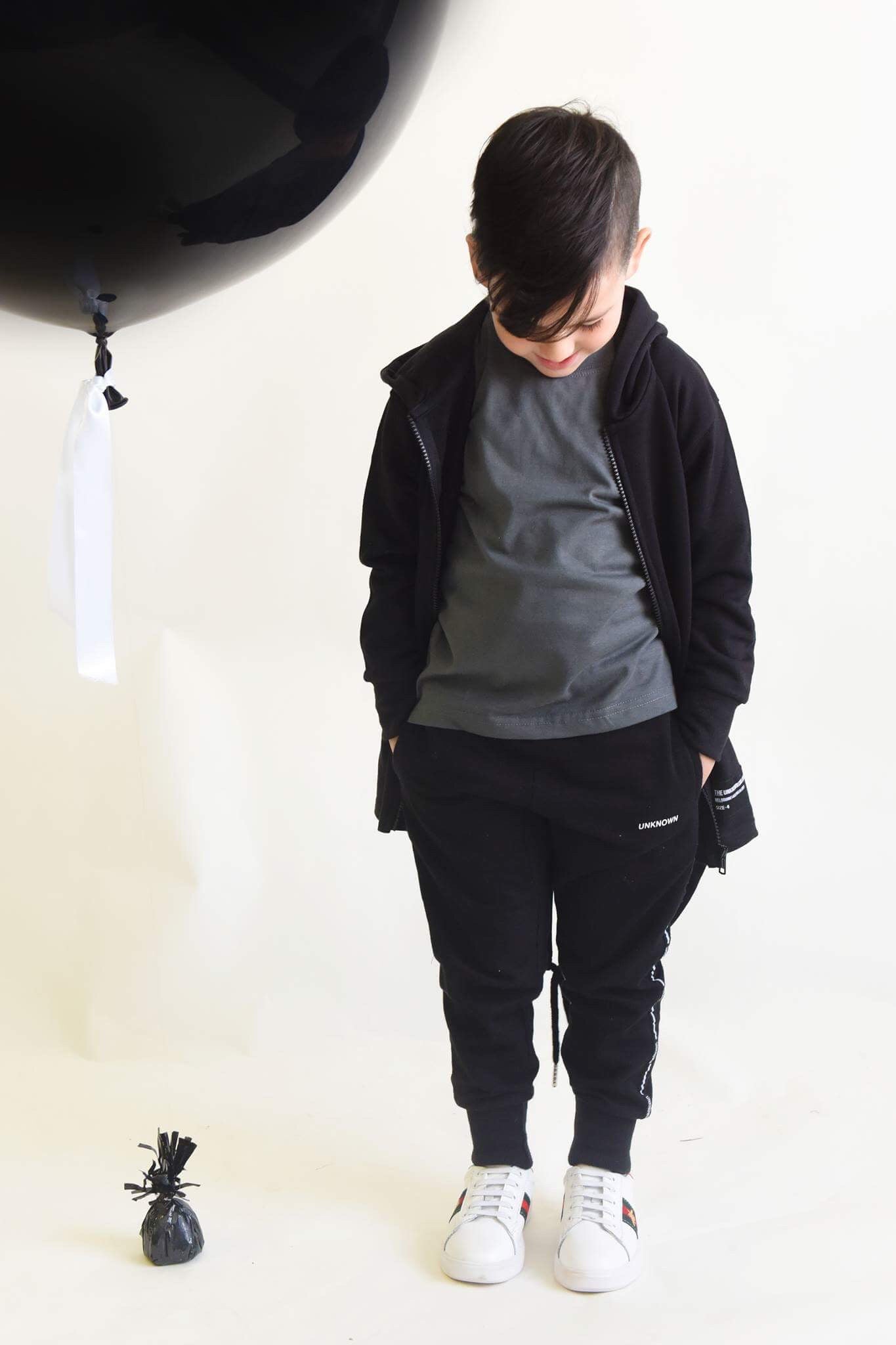Child wearing a black trench, gray t-shirt, and black "UNKNOWN" tracksuit pants, standing with hands in pockets, looking down. A large black balloon is seen in the background.