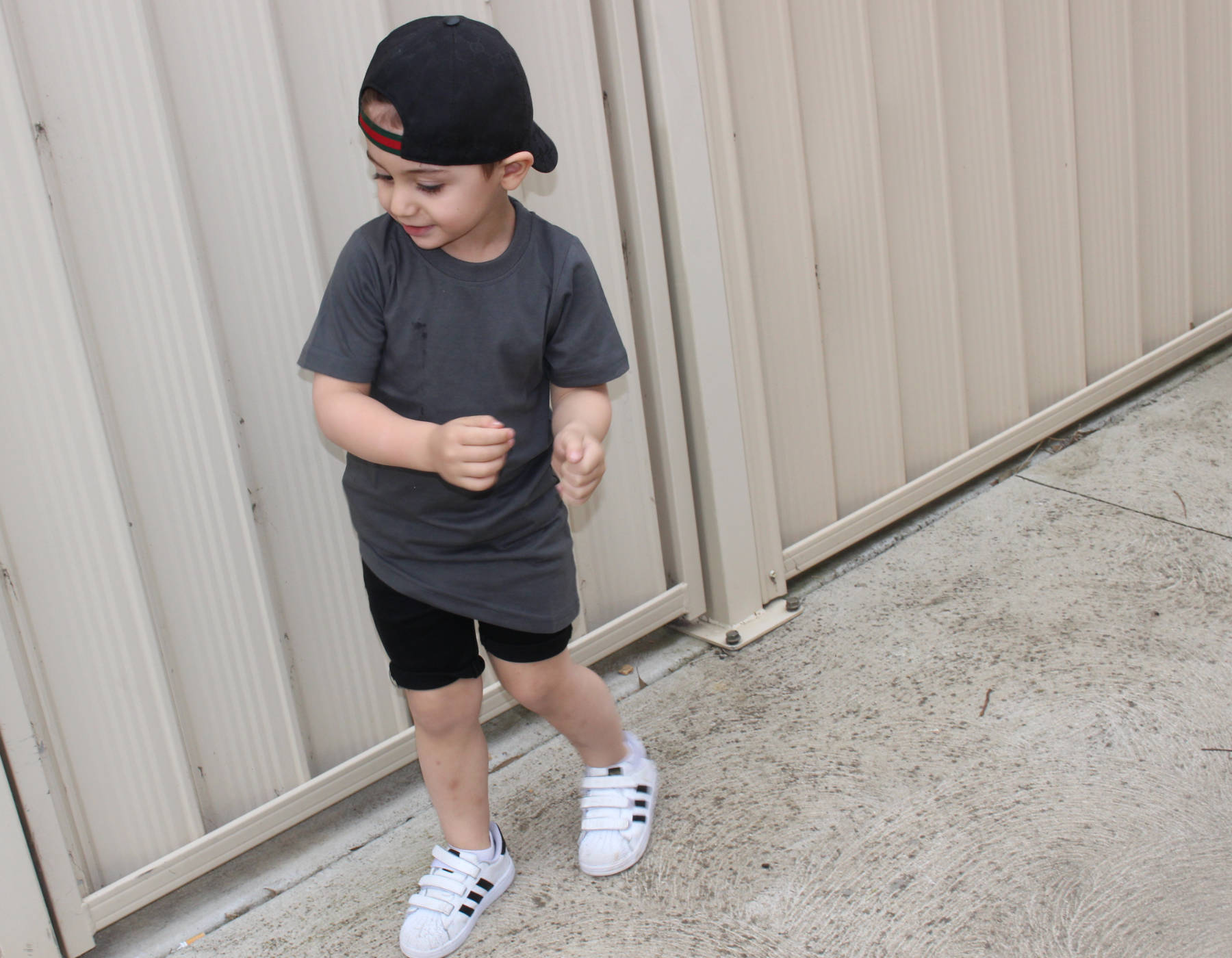 Child wearing a gray t-shirt, black shorts, and white sneakers standing against a beige wall.