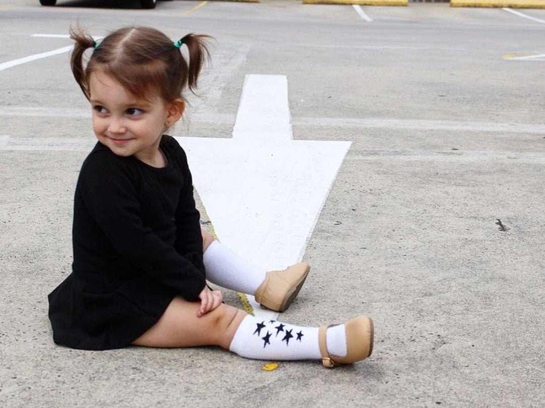 A young girl sitting on the ground in a parking lot, wearing THE UNKNOWN CHILDREN Classic Black Day Dress paired with white star-patterned knee-high socks and beige shoes. This 100% cotton dress is perfect for casual outings and playtime, combining comfort with a trendy look for active children.
