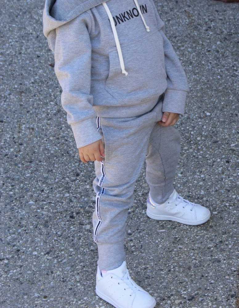 Child wearing a gray THE UNKNOWN CHILDREN tracksuit with white sneakers on a concrete surface. The pants feature a sleek design, perfect for active play and casual wear.