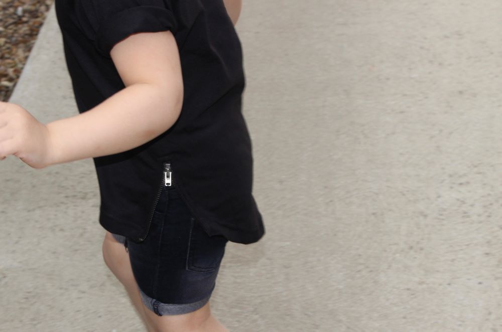Close-up of a child wearing THE UNKNOWN CHILDREN Zipped Curved Tee t-shirt in black, paired with denim shorts. The tee features a unique side zipper detail and curved hem, 
made from 100% superior cotton. 
Perfect for trendy and comfortable casual wear for kids. Ideal for active, fashion-forward children.