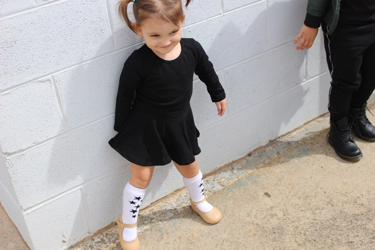A young girl wearing THE UNKNOWN CHILDREN Classic Black Day Dress, paired with white knee-high socks and beige shoes. This stylish, long-sleeve dress, made from 100% cotton, is perfect for both playtime and casual outings. Comfortable and trendy, it offers a versatile look for active kids.