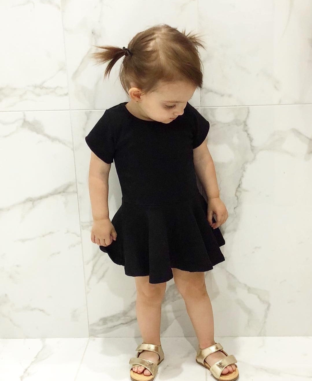 A young girl wearing THE UNKNOWN CHILDREN Classic Black Day Dress, paired with gold sandals. This short-sleeve dress, made from 100% cotton, offers a sleek and minimalist style, perfect for casual outings or special occasions. Comfortable and fashionable, it's a versatile wardrobe staple for active kids.