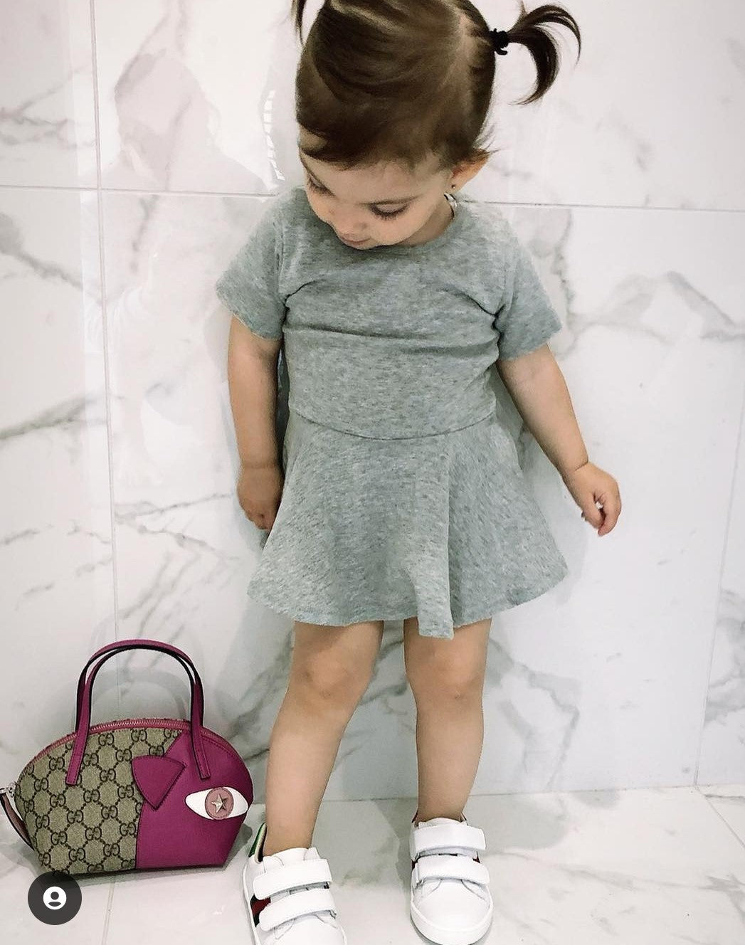 A young girl wearing THE UNKNOWN CHILDREN Classic Day Dress in gray, paired with white sneakers and standing next to a designer handbag. The dress, made from 100% cotton, offers a stylish and comfortable look, perfect for everyday wear. Ideal for active children who want both fashion and comfort.