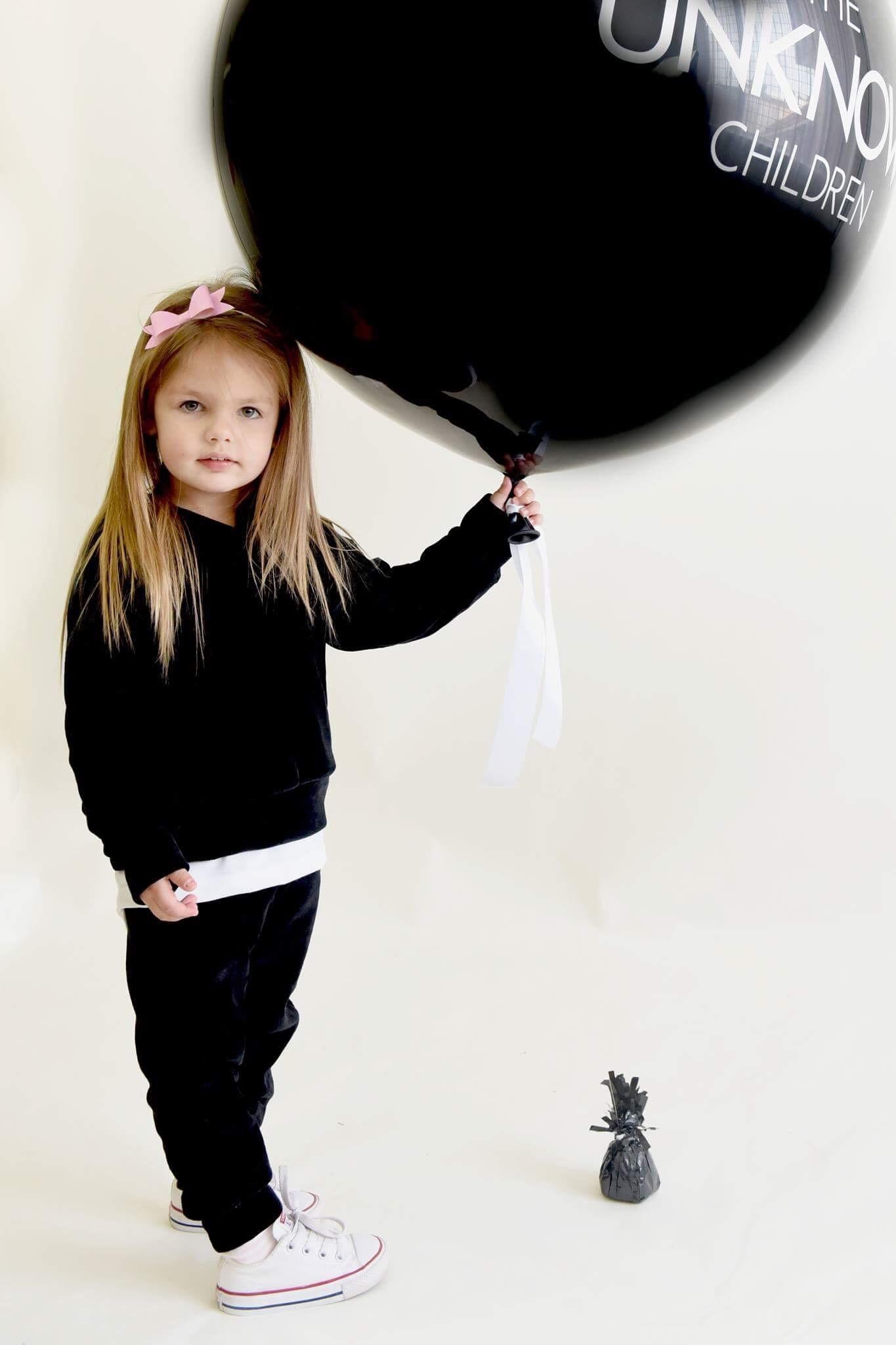 A young girl wearing THE UNKNOWN CHILDREN "CLASSICS" Black Velvet Set, standing and holding a large black balloon. The soft velvet outfit offers both comfort and style, paired with white sneakers, making it perfect for casual outings or playtime.
