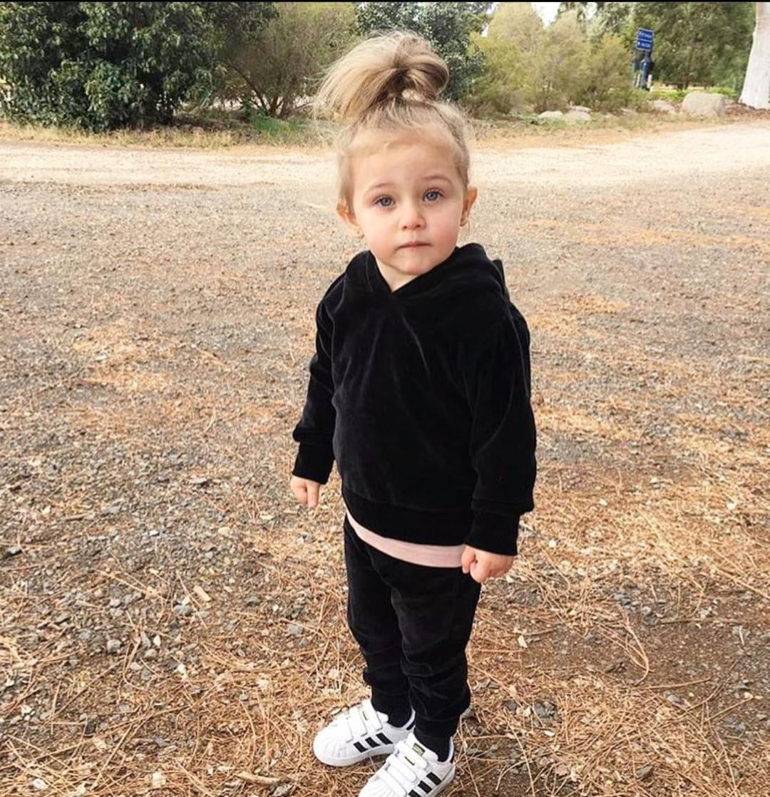 A young toddler wearing THE UNKNOWN CHILDREN "CLASSICS" Black Velvet Tracksuit and white sneakers, standing outdoors on a dirt path. The soft and stylish velvet set provides comfort and warmth, perfect for everyday wear.