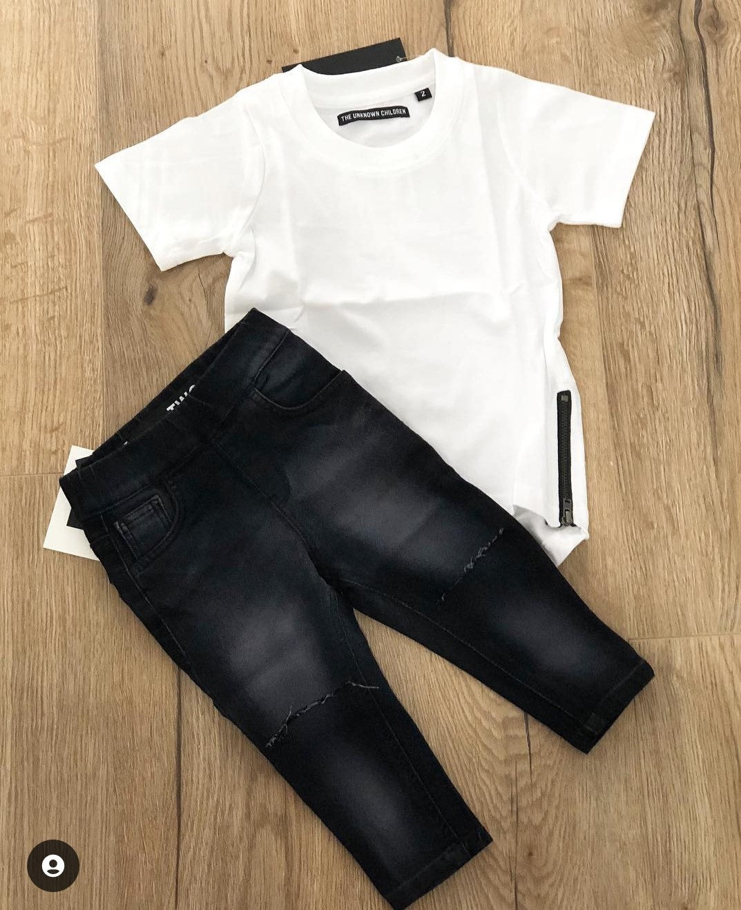 A stylish children's outfit from THE UNKNOWN CHILDREN collection featuring a simple white short-sleeve tee with a side zipper detail and a pair of black distressed jeans with subtle fade accents. The outfit is laid flat on a wooden floor, showcasing the modern and versatile design perfect for casual wear.