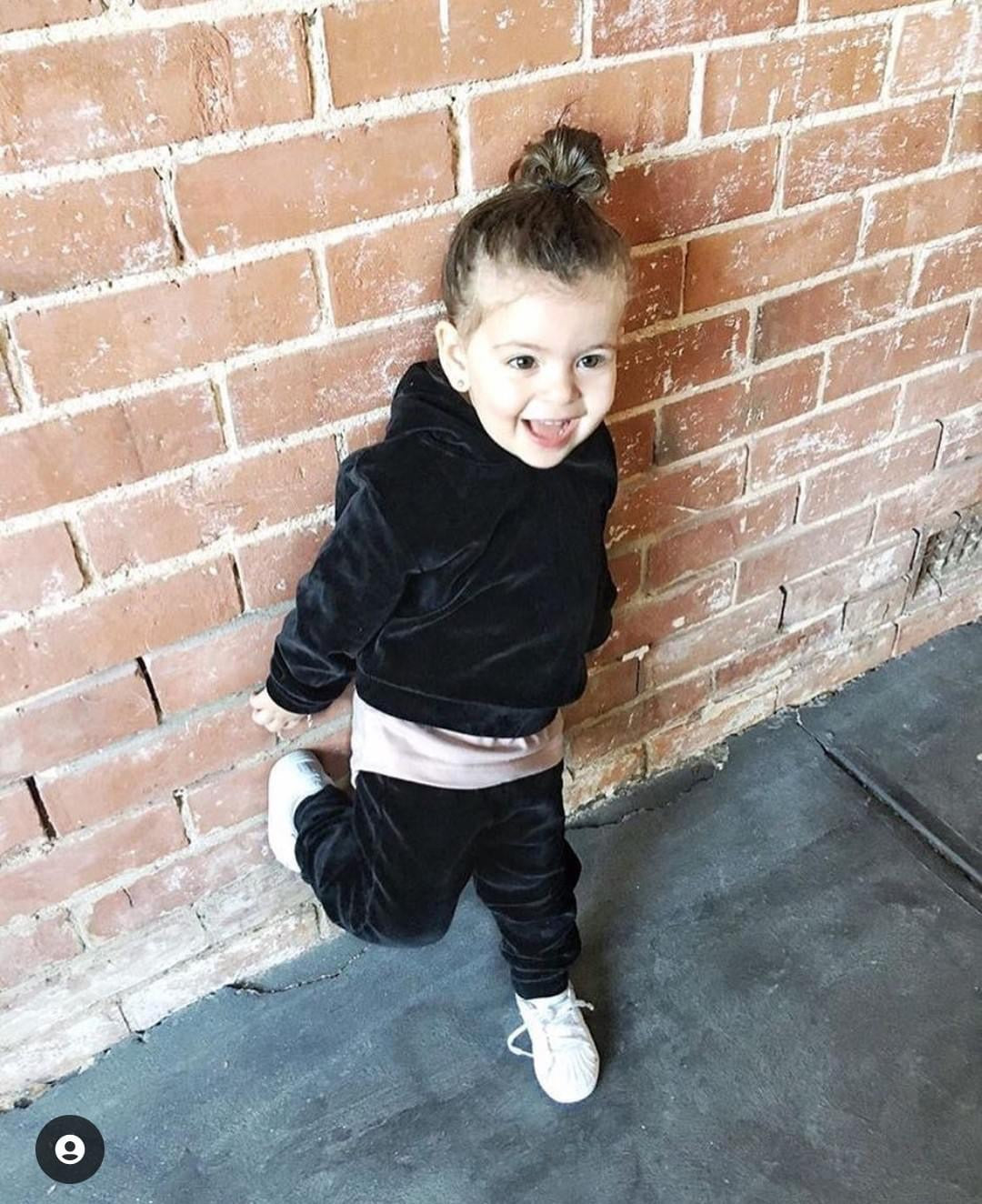 A happy toddler wearing THE UNKNOWN CHILDREN "CLASSICS" Black Velvet Tracksuit, standing against a rustic brick wall. The soft velvet set offers both comfort and style, paired with white sneakers for a cute and playful look.
