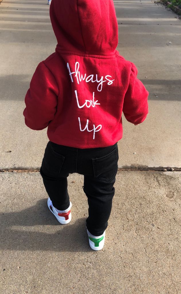A young child wearing THE UNKNOWN CHILDREN 'Originals' "Always Look Up" red hoodie walks on a concrete path. The unisex, limited-edition hoodie is made from 100% superior cotton, offering a trendy and comfortable style for boys and girls.