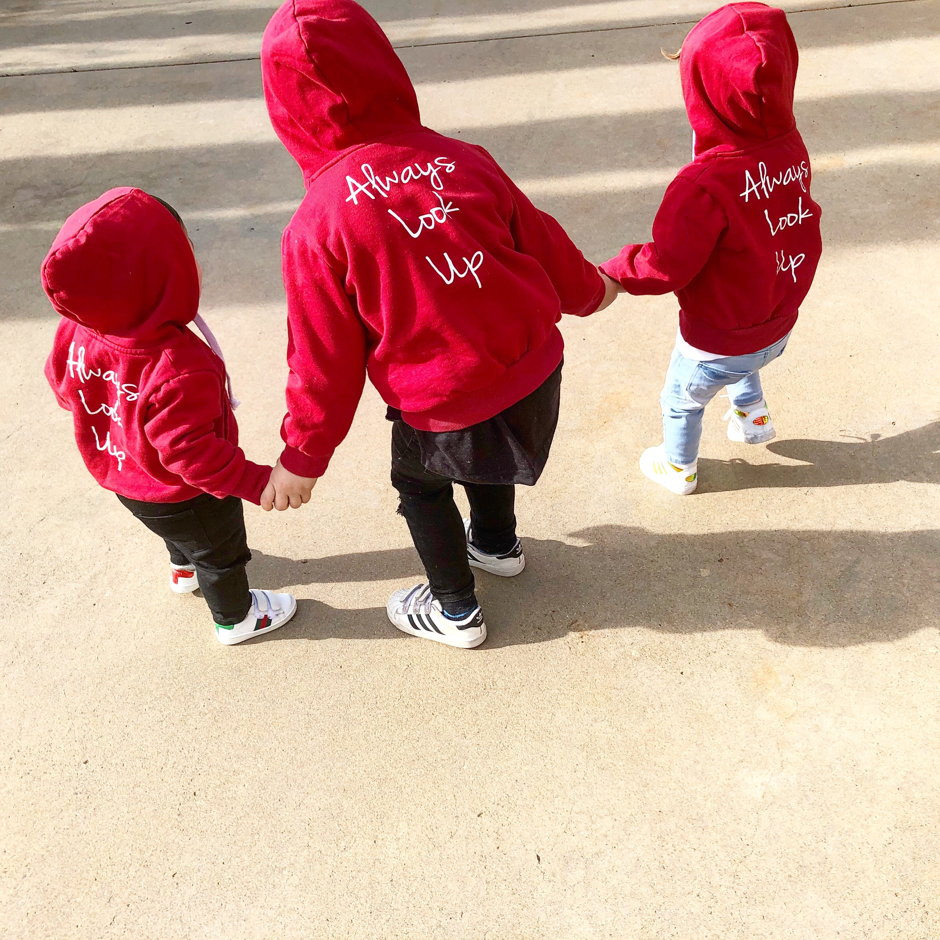 Three children wearing THE UNKNOWN CHILDREN 'Originals' "Always Look Up" red hoodies, walking hand in hand. This unisex, limited-edition hoodie is made from 100% superior cotton, showcasing a trendy and comfortable design for boys and girls.
