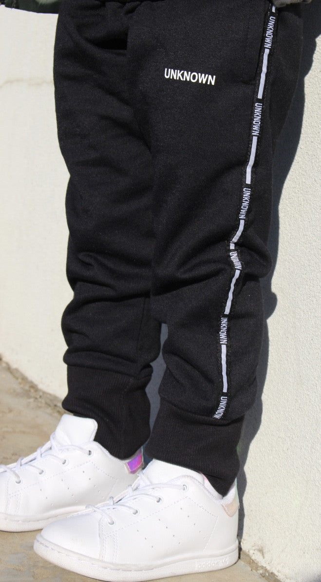 Close-up of a child wearing THE UNKNOWN CHILDREN black tracksuit pants with the word "UNKNOWN" printed along the side seam, paired with white sneakers. The pants feature a sleek design, perfect for active play and casual wear.