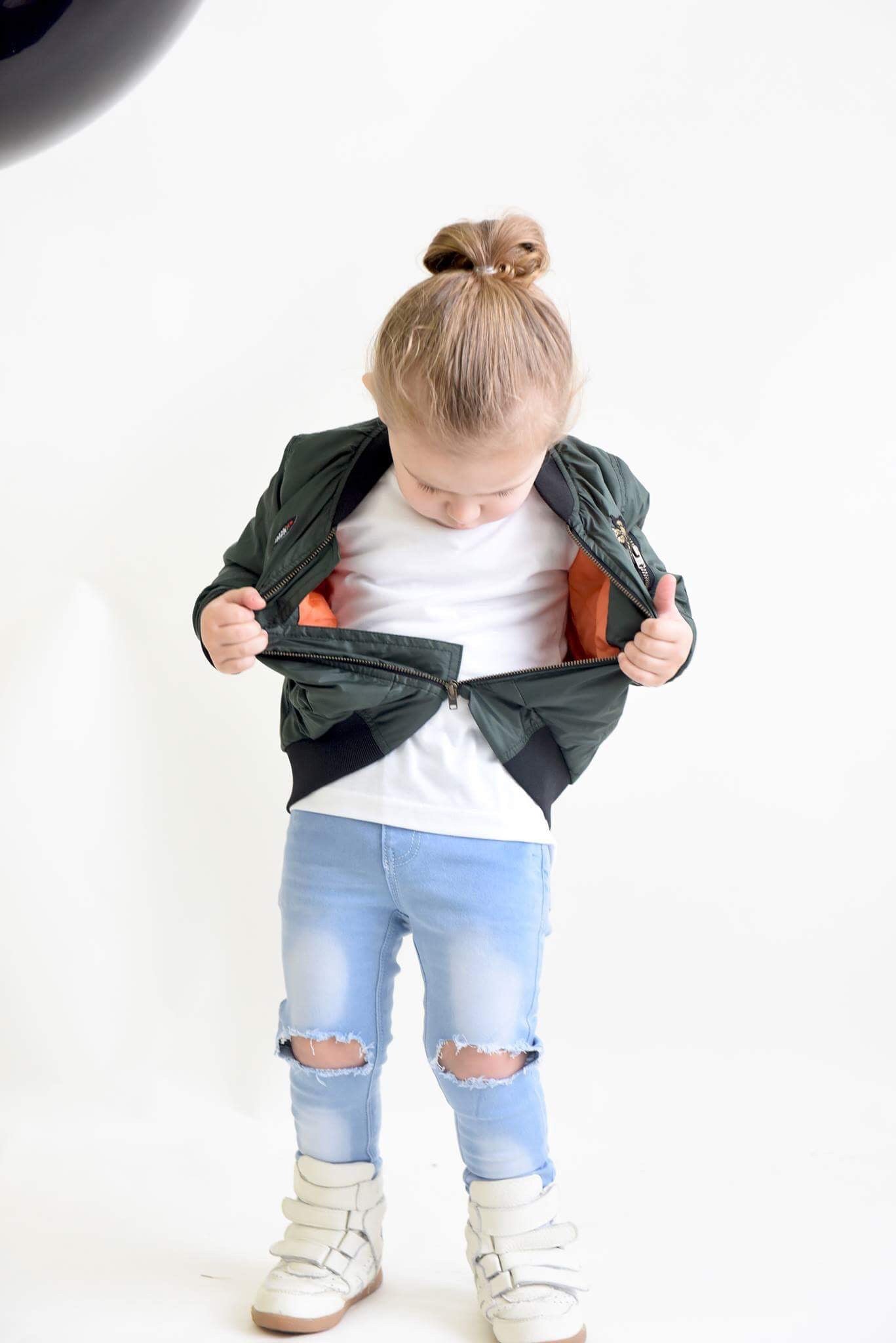 A young child wearing a green bomber jacket with an orange inner lining, a white T-shirt, and light blue ripped jeans from THE UNKNOWN CHILDREN collection. The child is looking down while unzipping the jacket, revealing the bright inner lining. The outfit is completed with white high-top sneakers. The photo is taken in a bright studio with a clean, white background.