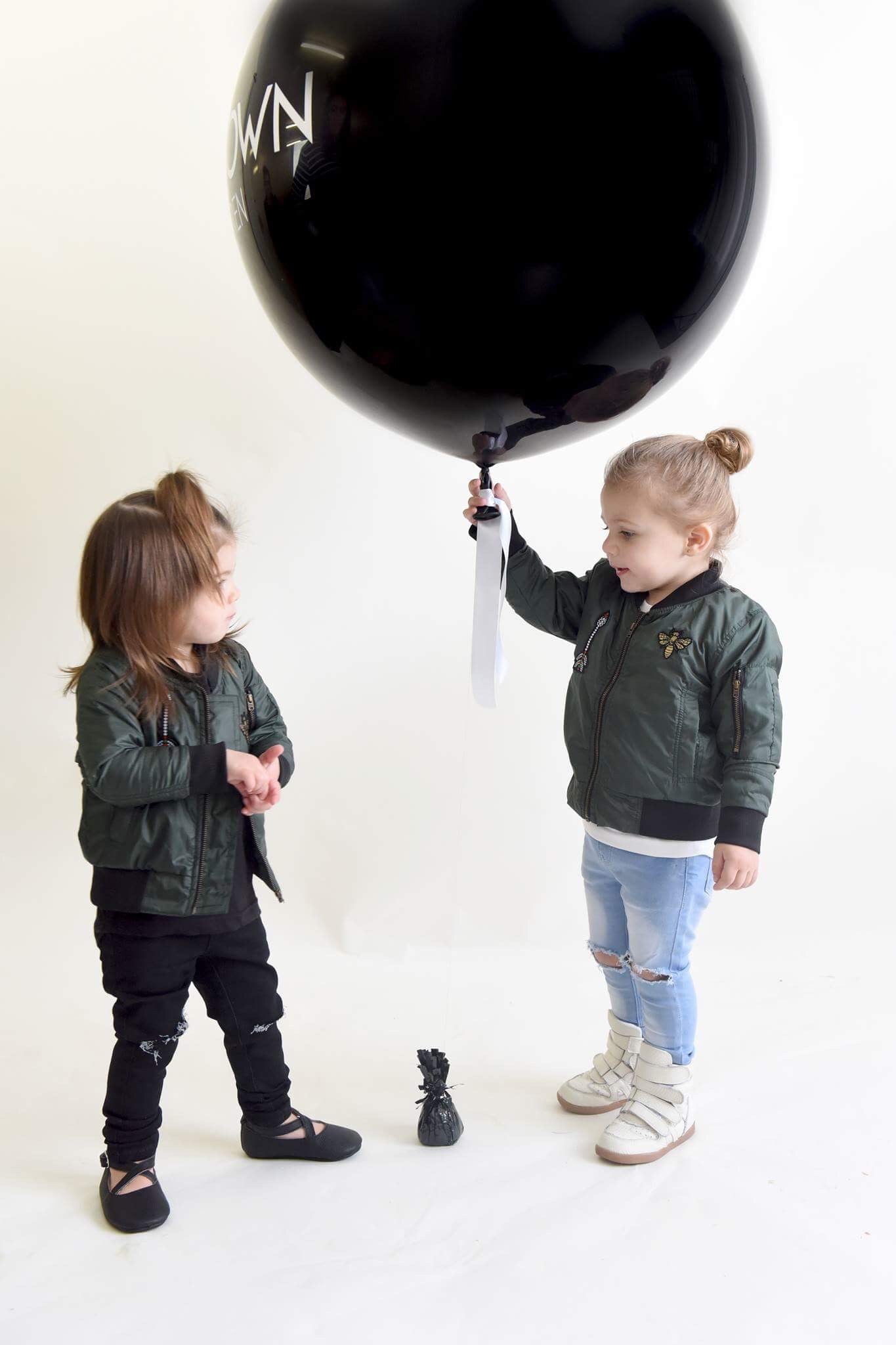 Two young children wearing matching green bomber jackets from THE UNKNOWN CHILDREN Originals collection. One child holds a large black balloon, while the other looks on. One child wears THE UNKNOWN CHILDREN light blue ripped jeans and white high-top sneakers, while the other is dressed in THE UNKNOWN CHILDREN black JEANS and black shoes. The setting is a bright studio with a minimalistic background.