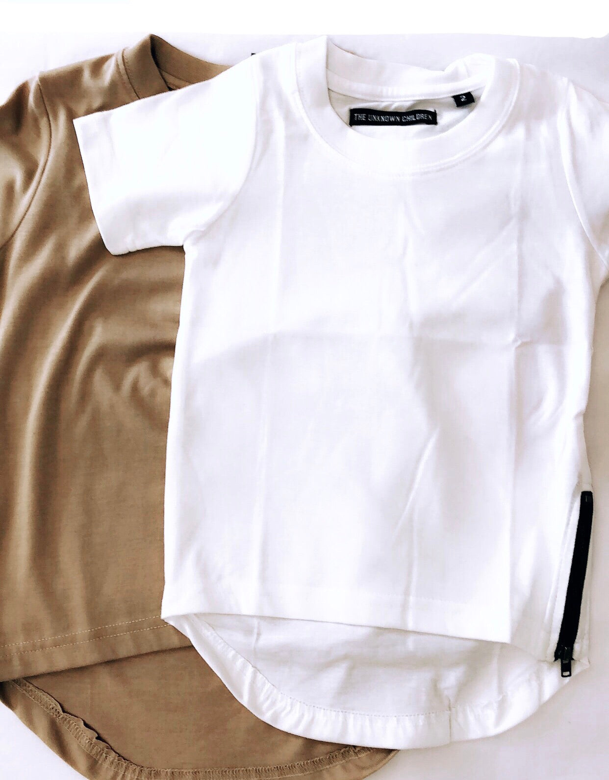 Two Zipped Curved Tees from THE UNKNOWN CHILDREN, featuring a beige and white option, both made from 100% superior cotton. These stylish and comfortable tees have a unique zip detail and curved hem, perfect for adding a modern touch to any child's wardrobe. Ideal for trendy, fashion-forward kids.
