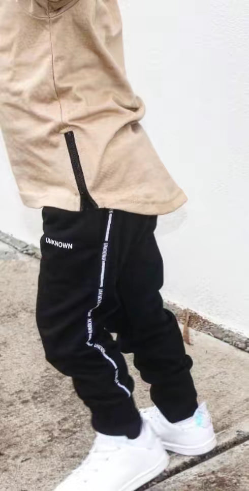 Child wearing a beige curved hem tee with a side zipper detail, paired with black "UNKNOWN" tracksuit pants and white sneakers. The outfit combines comfort and style, perfect for casual everyday wear.