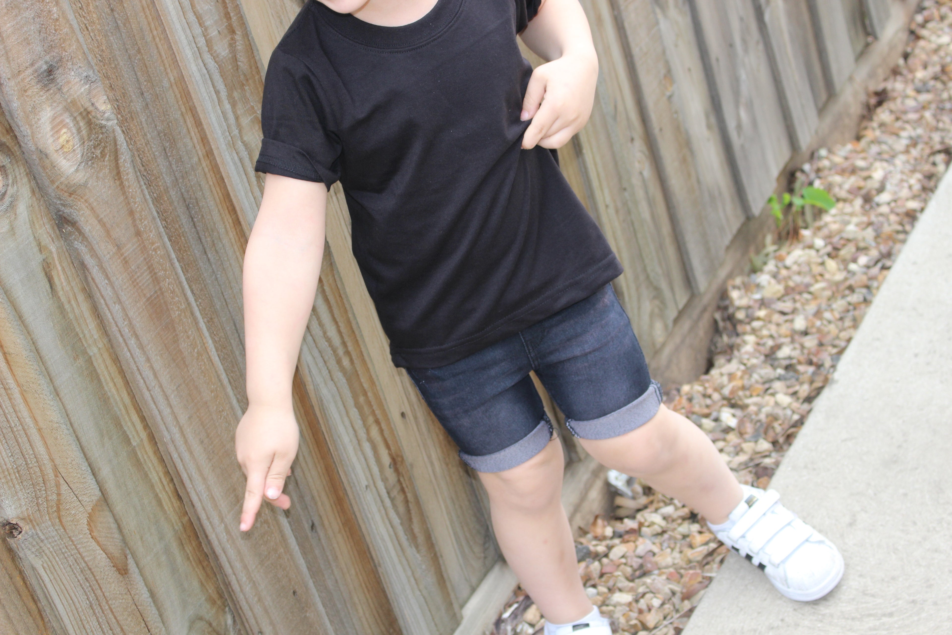 Child wearing black stretch denim shorts and a black t-shirt, paired with white sneakers, standing near a wooden fence. The denim shorts feature rolled hems, offering both comfort and durability for everyday play.