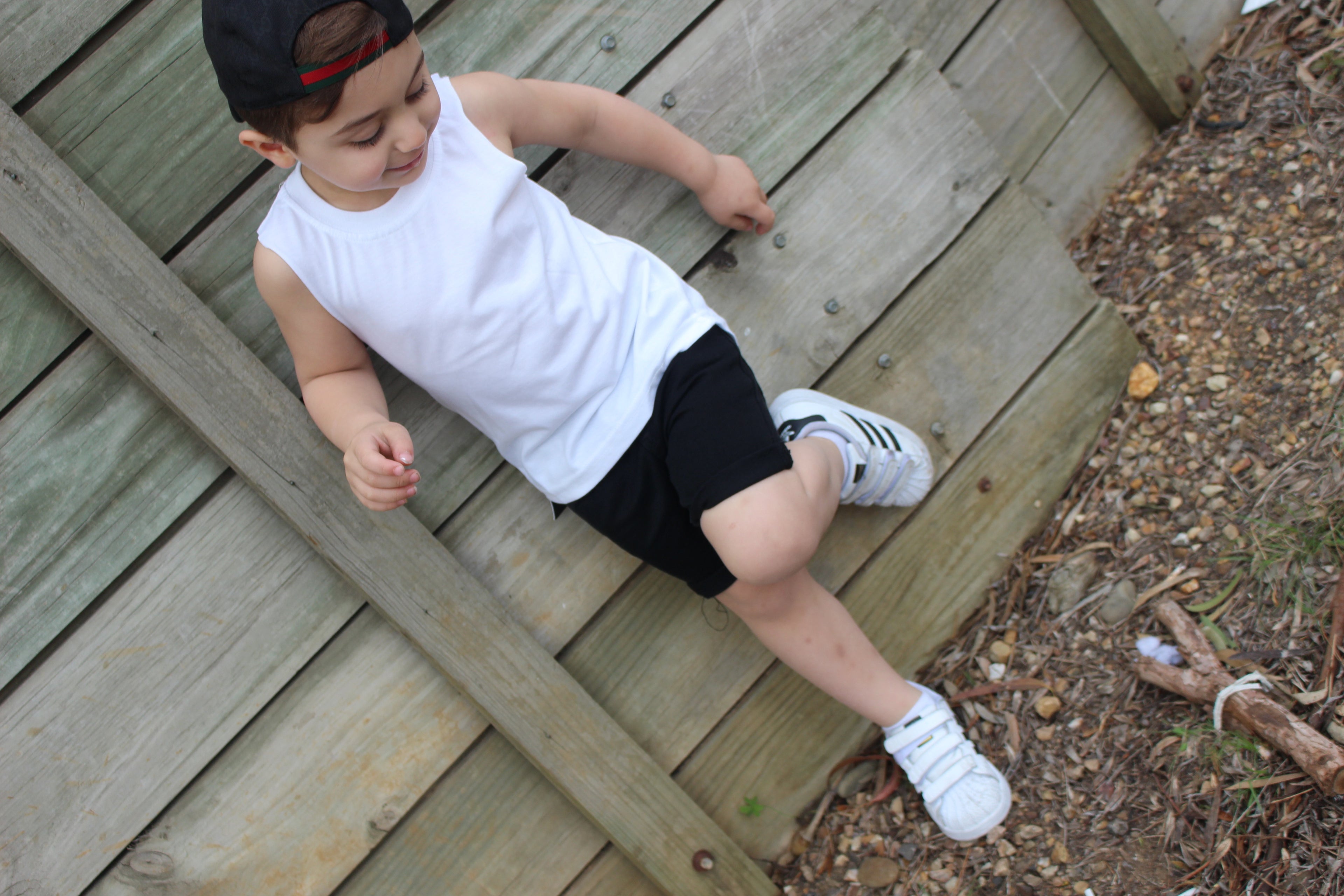 Child wearing a white sleeveless shirt, black shorts, and white sneakers, relaxing against a wooden fence. The casual summer outfit is perfect for outdoor play, paired with a backward cap for a trendy look.