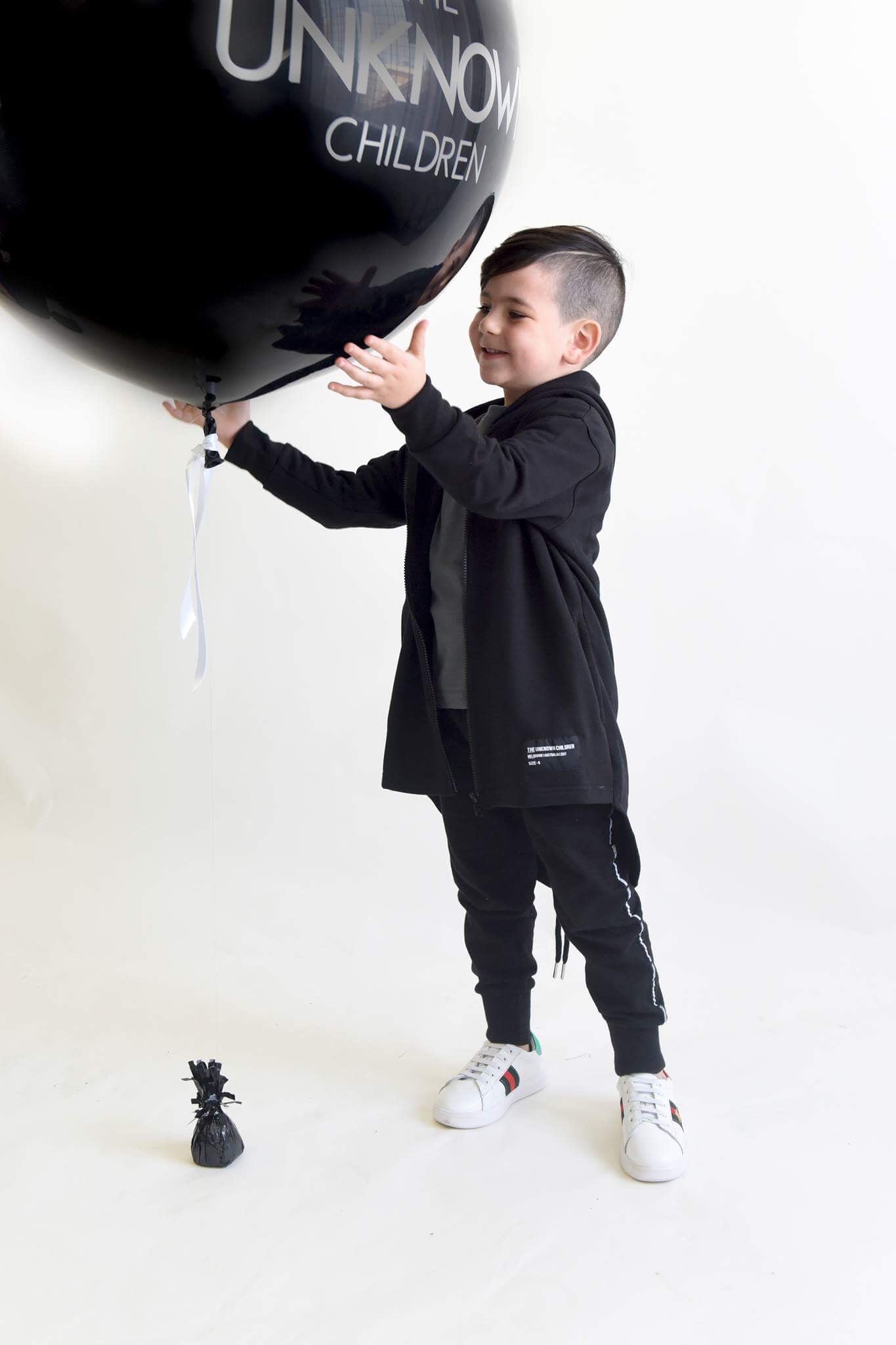 Child smiling and reaching out to hold a large black balloon with "THE UNKNOWN CHILDREN" logo, wearing a black trench and tracksuit pants with white sneakers. The child is playing in a minimalist setting with a small black decorative item on the floor.