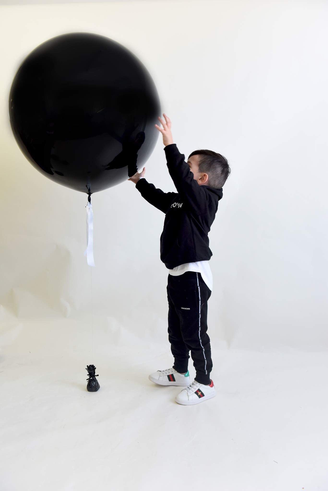 A young boy smiling and playing with a large black balloon branded "The Unknown Children." He is wearing a black hoodie, black joggers, and white sneakers, creating a stylish and comfortable look for casual wear.