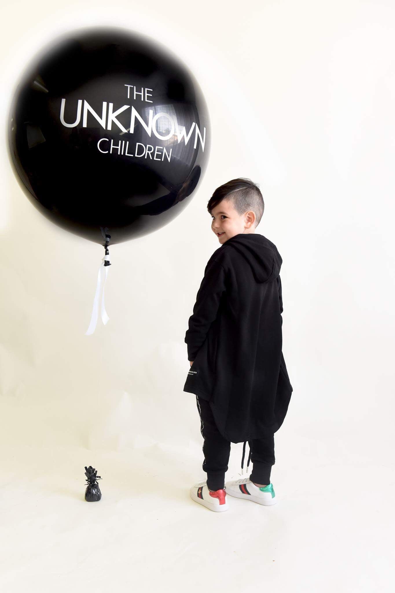Child wearing a black trench and tracksuit from THE UNKNOWN CHILDREN, standing sideways with a smile. A large black balloon with "THE UNKNOWN CHILDREN" branding hovers next to the child. White sneakers with red and green accents complete the look.