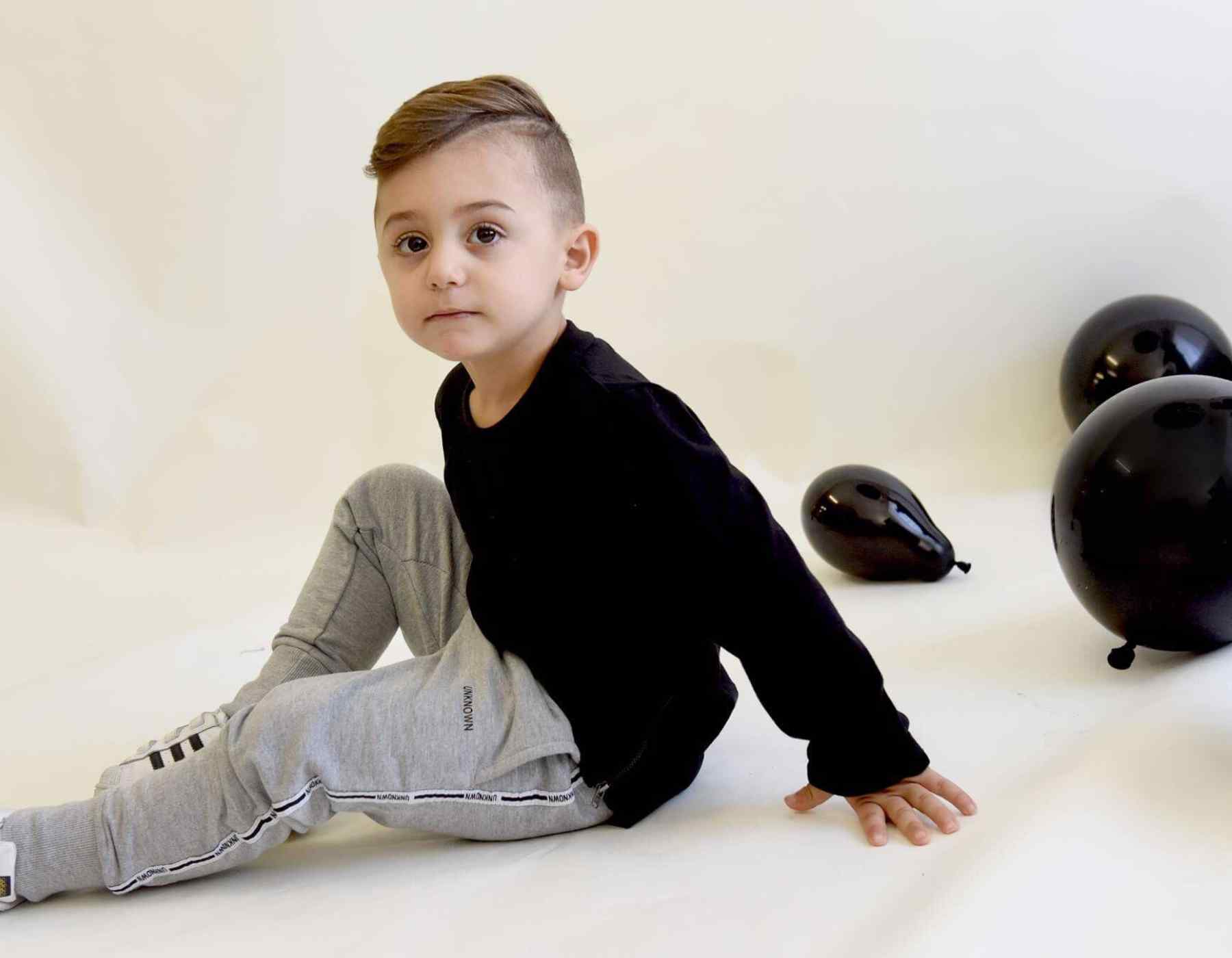Child in black shirt and gray pants sitting on a white surface with black balloons.