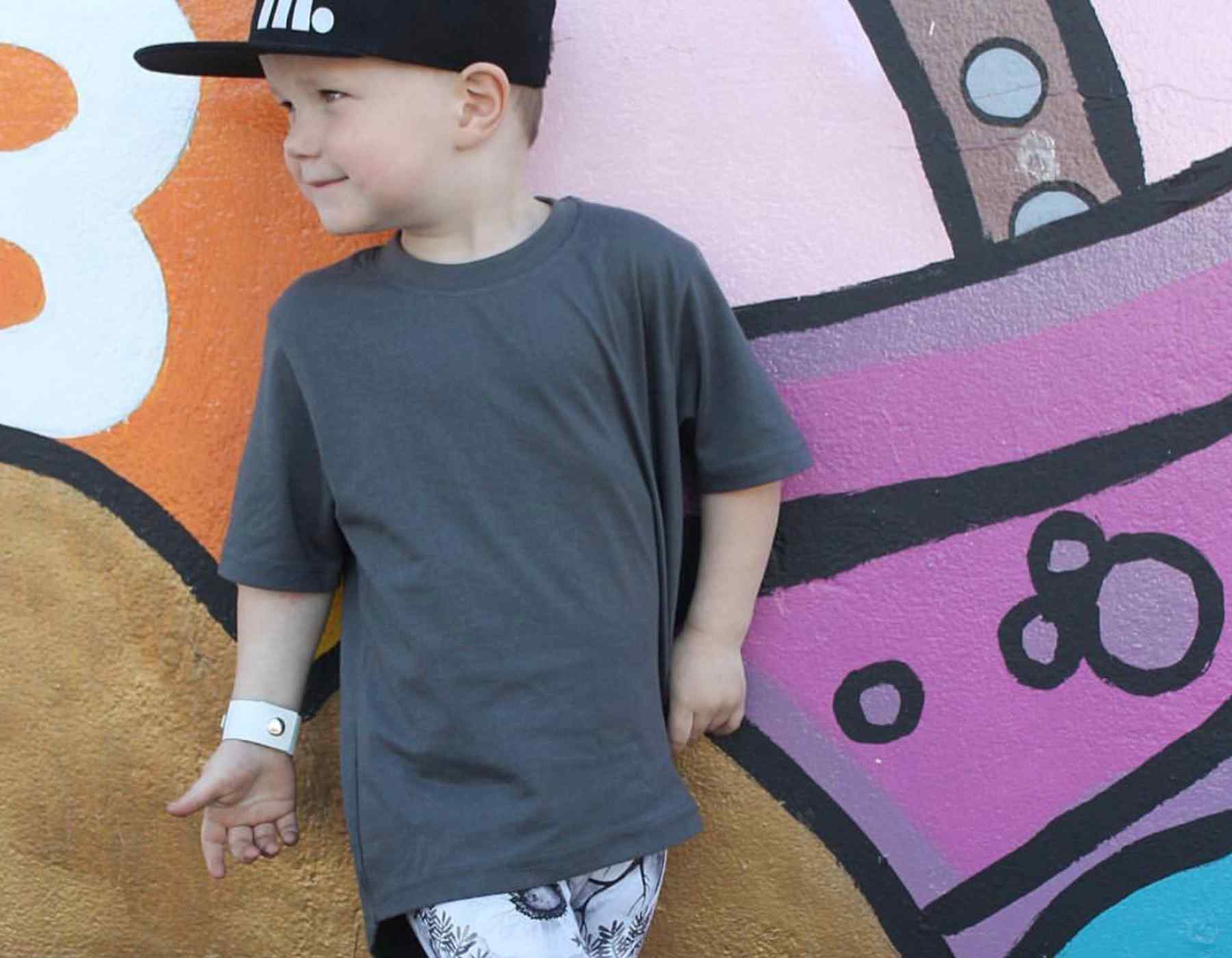 Child wearing a dark gray t-shirt and black cap in front of a colorful mural.